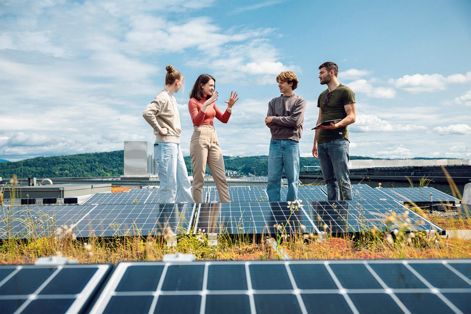 Vier junge Erwachsene stehen und unterhalten sich auf einem Dach mit Sonnenkollektoren, umgeben von Grün und einer malerischen Landschaft im Hintergrund unter einem blauen Himmel mit Wolken.