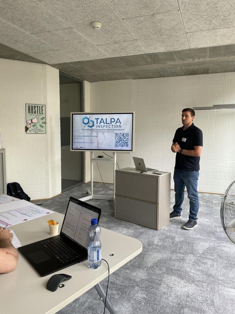 Ein Mann steht neben einem Fernsehbildschirm und hält eine Präsentation mit dem Titel TALPA INSPECTION in einem modernen Büro. Zwei Personen sitzen an einem Tisch, an dem im Vordergrund Laptops, Notebooks und eine Wasserflasche zu sehen sind.