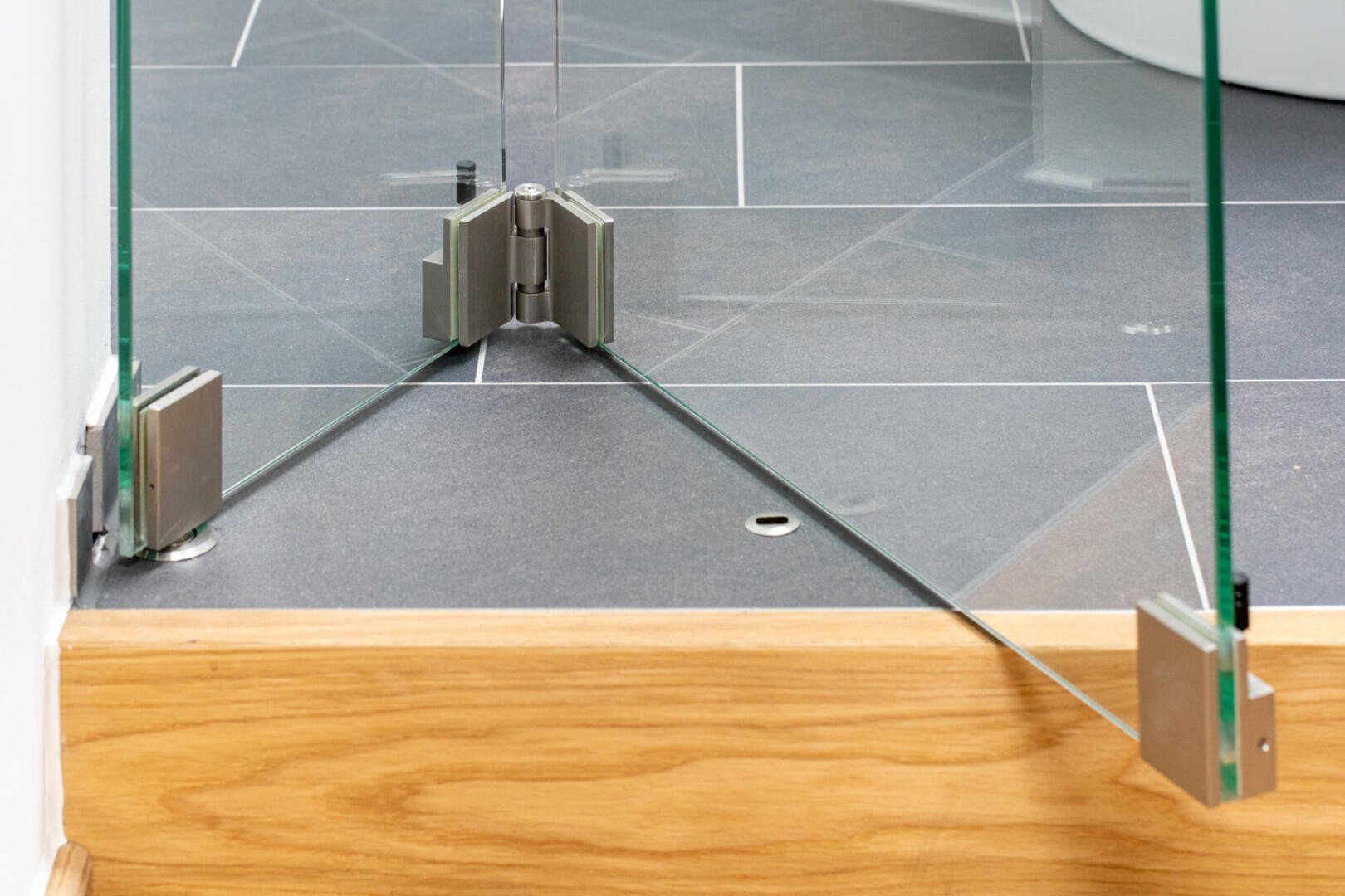 Nahaufnahme einer rahmenlosen Glasduschtür mit Metallscharnieren, die auf einem gefliesten Badezimmerboden neben einer Holzstufe installiert ist. Die Glaspaneele bilden am Boden eine Ecke.