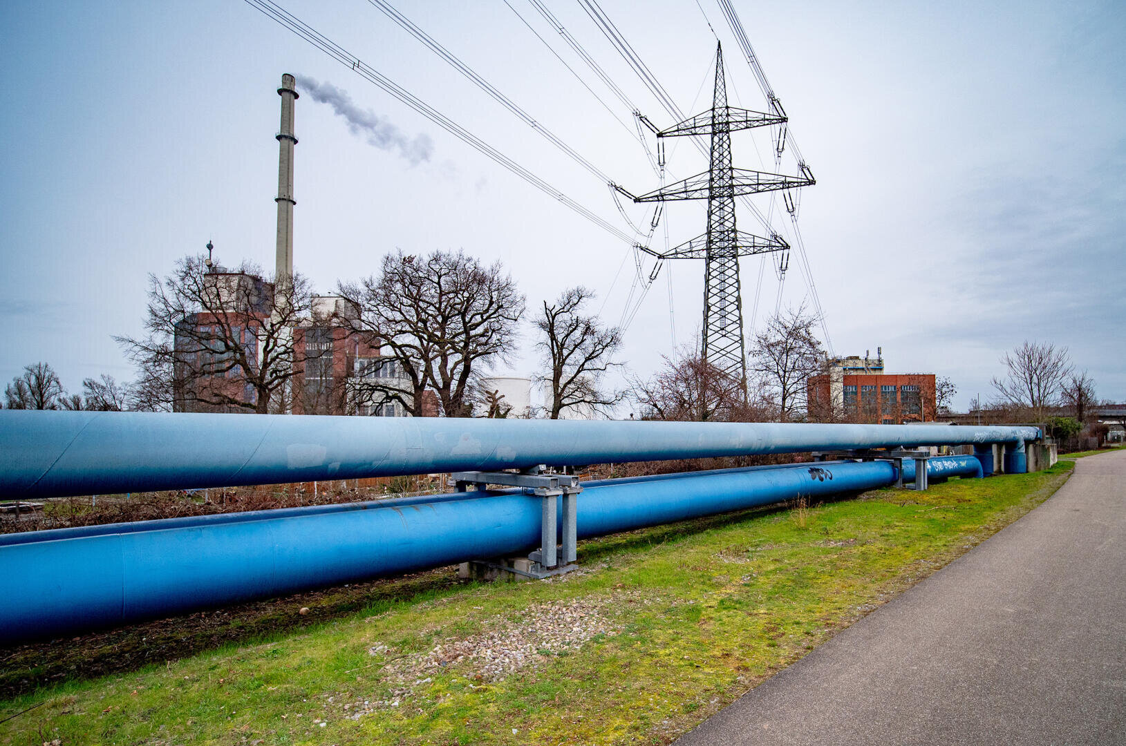 Große blaue Industrierohre verlaufen parallel zu einem gepflasterten Weg, während im Hintergrund ein Hochspannungsmast und eine Fabrik Rauch ausstoßen. Unter einem bewölkten Himmel sind Bäume ohne Blätter und Grasflächen zu sehen.
