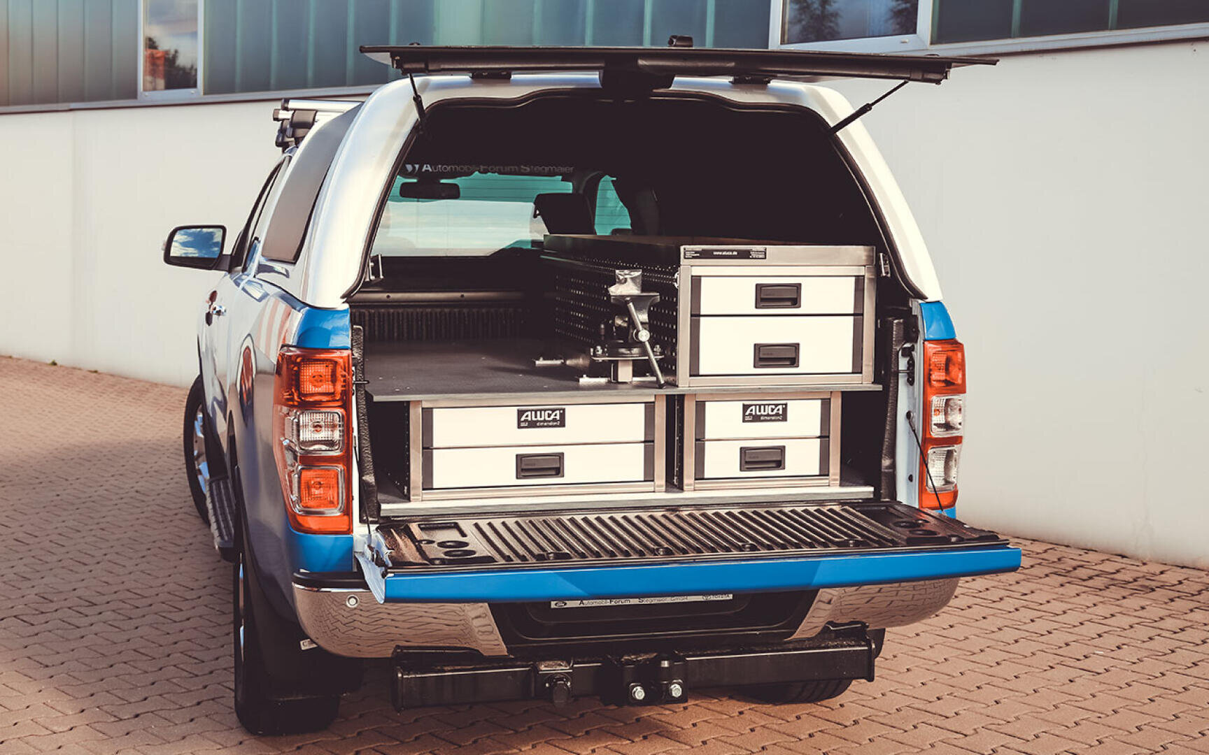Ein blau-weißer Pickup mit geöffneter Heckklappe und offenem Heckverdeck, das ein geordnetes Aufbewahrungssystem mit Metallschubladen und -fächern offenbart, die ordentlich in die Ladefläche eingebaut sind.