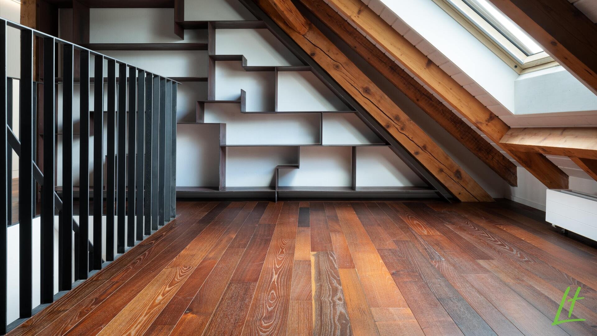 Une pièce mansardée moderne avec des poutres en bois inclinées, un puits de lumière, un sol en bois sombre et une bibliothèque géométrique unique intégrée dans le mur en angle. Une rampe en métal noir est visible sur le côté gauche.