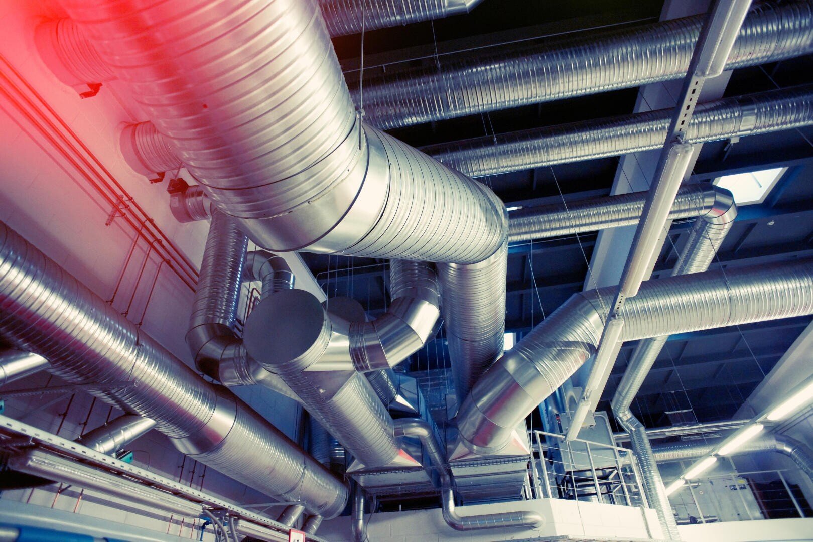 Große silberne Lüftungskanäle und -rohre verlaufen an der Decke einer Industrieanlage, reflektieren das Licht und bilden ein komplexes Netzwerk, in dem in der oberen linken Ecke rotes Licht aufflackert.