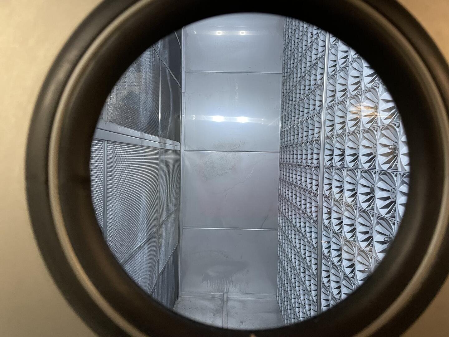 Blick durch ein rundes Fenster in einen metallischen, industriellen Raum mit Maschen und gemusterten Gittern an den Wänden, beleuchtet von einer Leuchtstoffröhre.