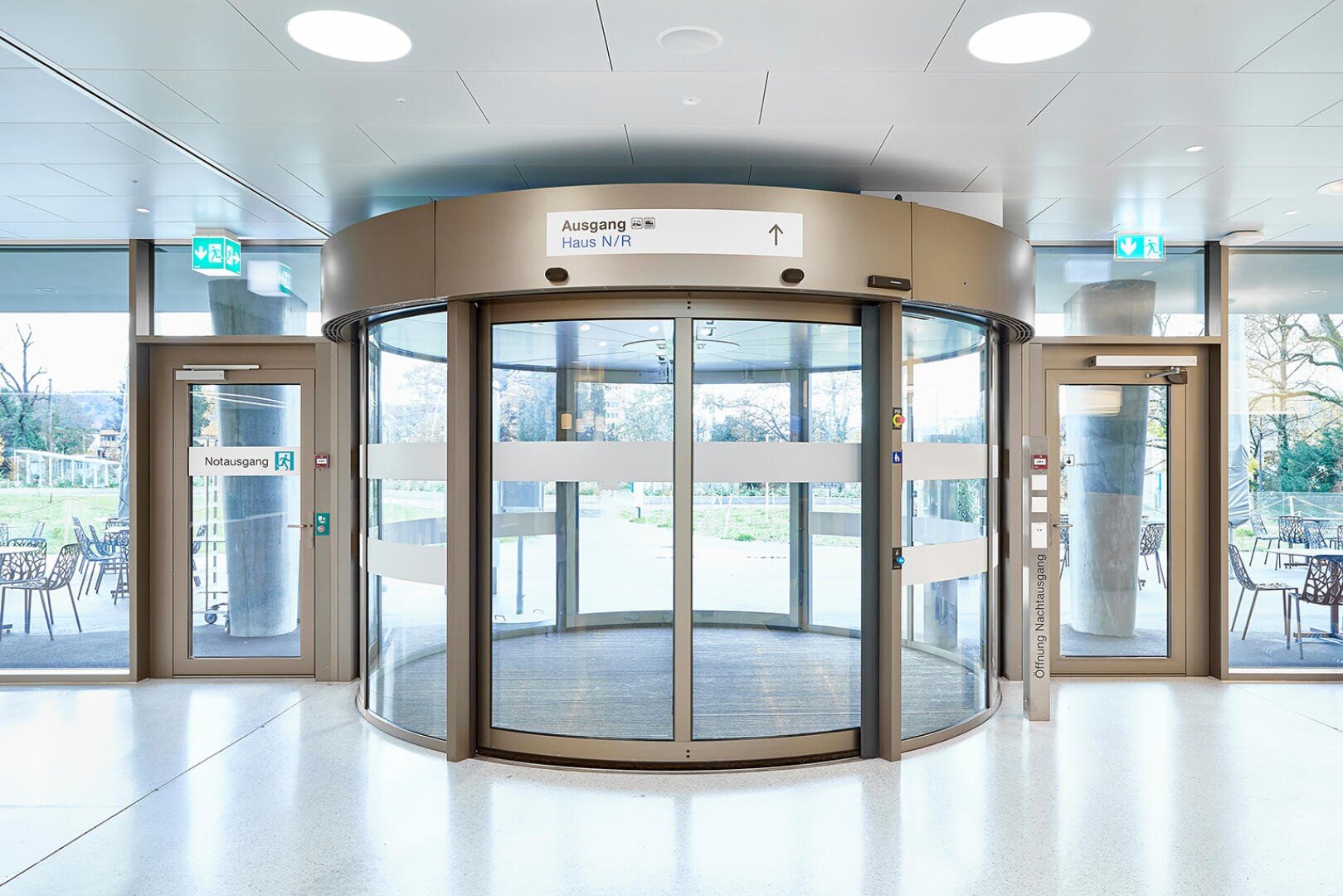 Ein moderner Gebäudeeingang mit einer großen Glasdrehtür in der Mitte, flankiert von zwei Glasausgangstüren. Schilder über den Türen weisen den Weg in deutscher Sprache. Draußen sind Terrassenmöbel und Grünflächen zu sehen.