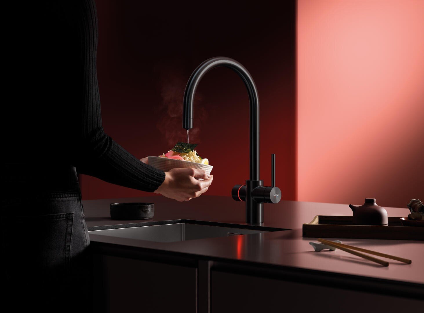 Eine Person hält eine dampfende Schüssel unter einem modernen schwarzen Küchenwasserhahn auf einer dunklen Arbeitsplatte, mit minimalistischem Dekor und einem warmen roten Licht im Hintergrund.