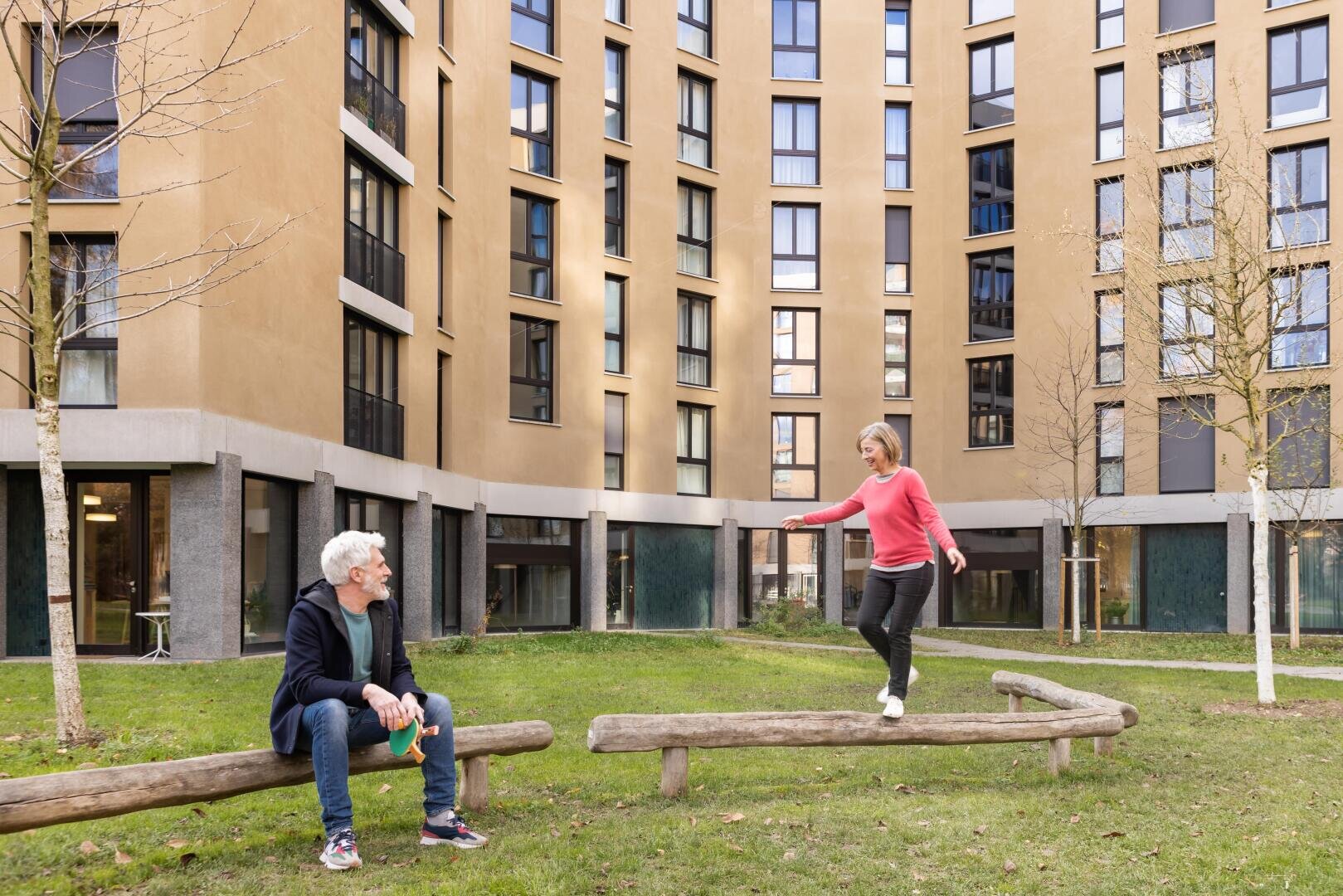 Eine Frau balanciert auf einem Holzbalken, während ein Mann in der Nähe sitzt und sie beobachtet. Beide befinden sich in einem grasbewachsenen Innenhof, der von einem modernen, mehrstöckigen Wohnhaus umgeben ist.