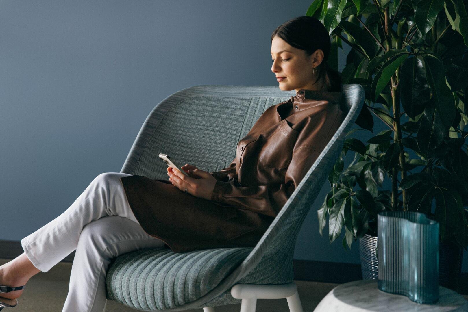Eine Frau sitzt in einem modernen Stuhl und schaut auf ihr Telefon. Sie trägt einen braunen Mantel und eine weiße Hose. Neben ihr steht eine große Grünpflanze und im Vordergrund steht eine blaue Vase auf einem runden Tisch. Die Wand ist blau gestrichen.