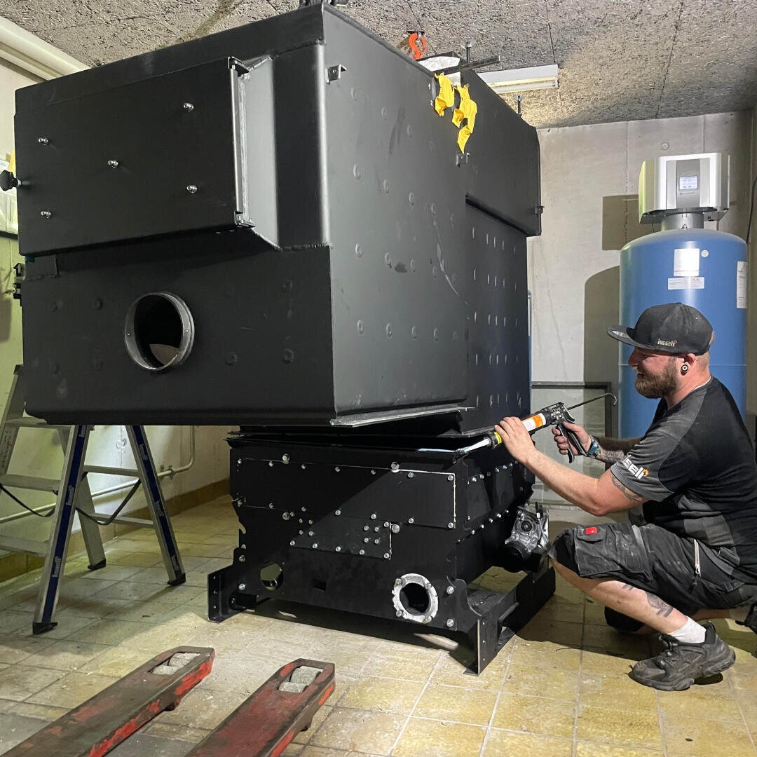 Ein Mann in Arbeitskleidung benutzt ein Werkzeug, um eine große, schwarze Industriemaschine in einem gefliesten Raum mit verschiedenen Geräten und einem Wassererhitzer im Hintergrund zu montieren oder zu reparieren.