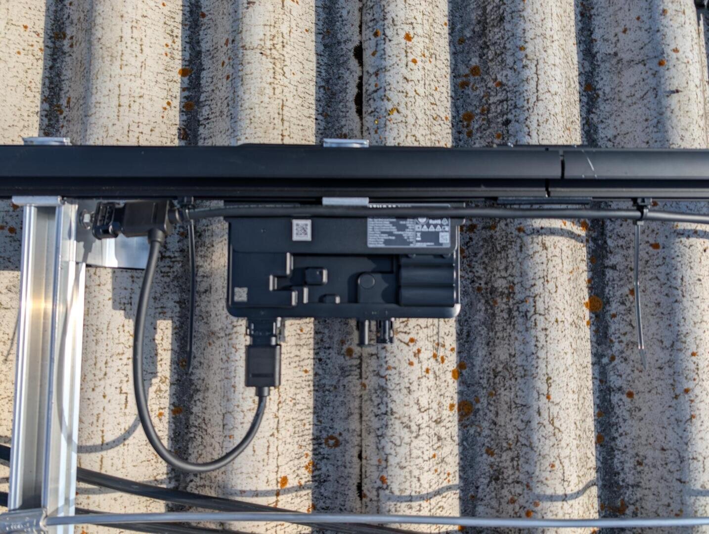 Nahaufnahme eines schwarzen elektrischen Geräts, das auf einem Metallrahmen auf einem gewellten, verwitterten Dach mit Flechtenflecken montiert ist. Die Kabel sind an das Gerät angeschlossen.