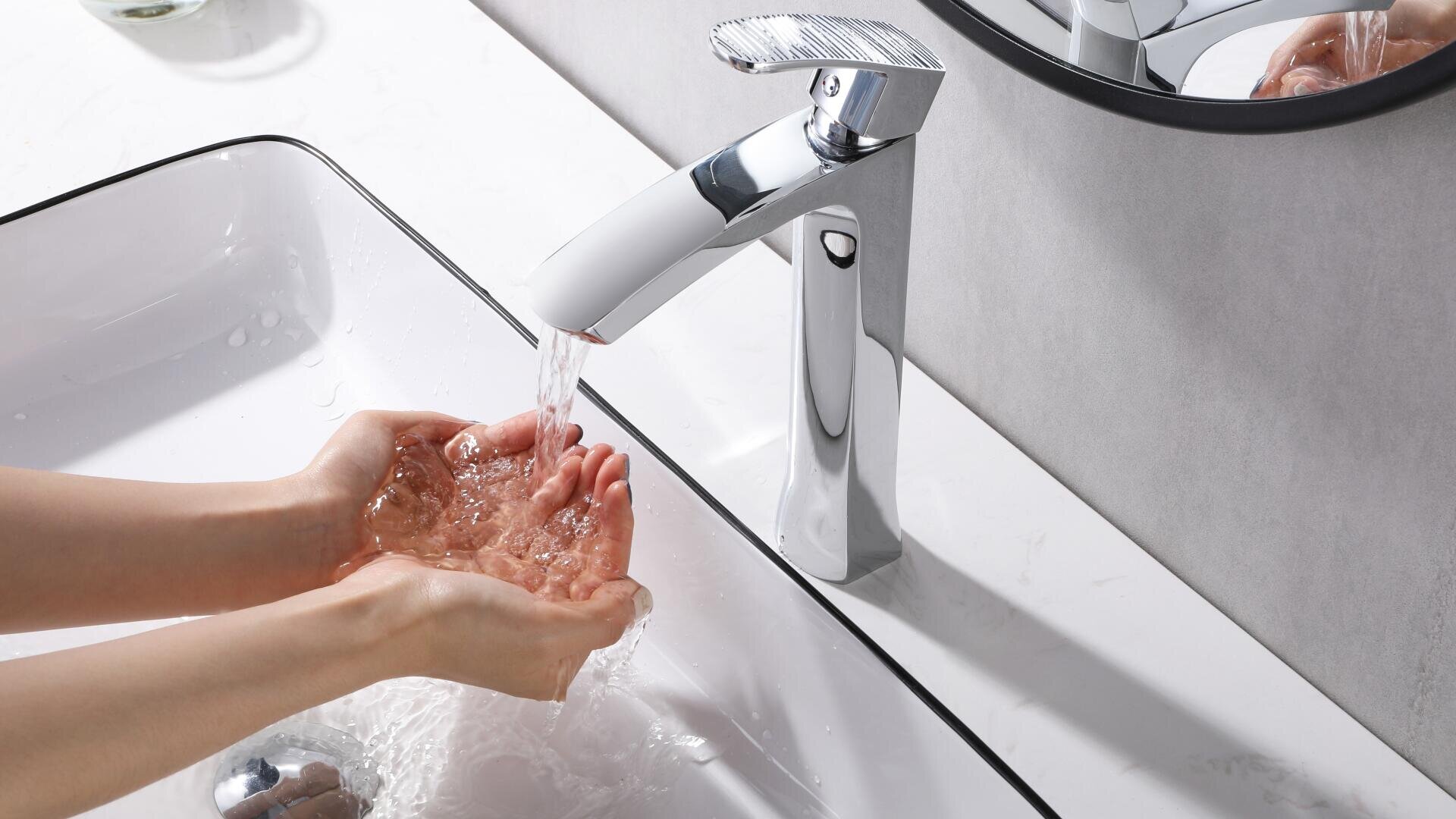 Eine Person wäscht sich die Hände unter einem modernen verchromten Wasserhahn in einem weißen Waschbecken, wobei das Wasser spritzt. Ein runder Spiegel, ein Seifenspender und eine Glaskerze stehen auf der weißen Arbeitsplatte.