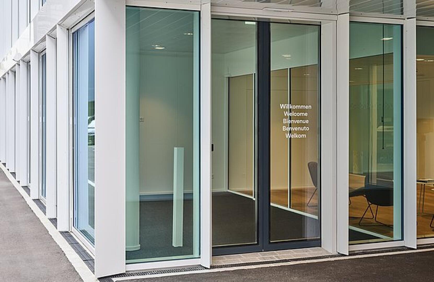 Gläserne Eingangstür zu einem modernen Gebäude mit der Aufschrift "Willkommen, Welcome, Bienvenue, Benvenuto, Welkom" in weißen Buchstaben und einem Stuhl, der in der Nähe des Holzbodens zu sehen ist.