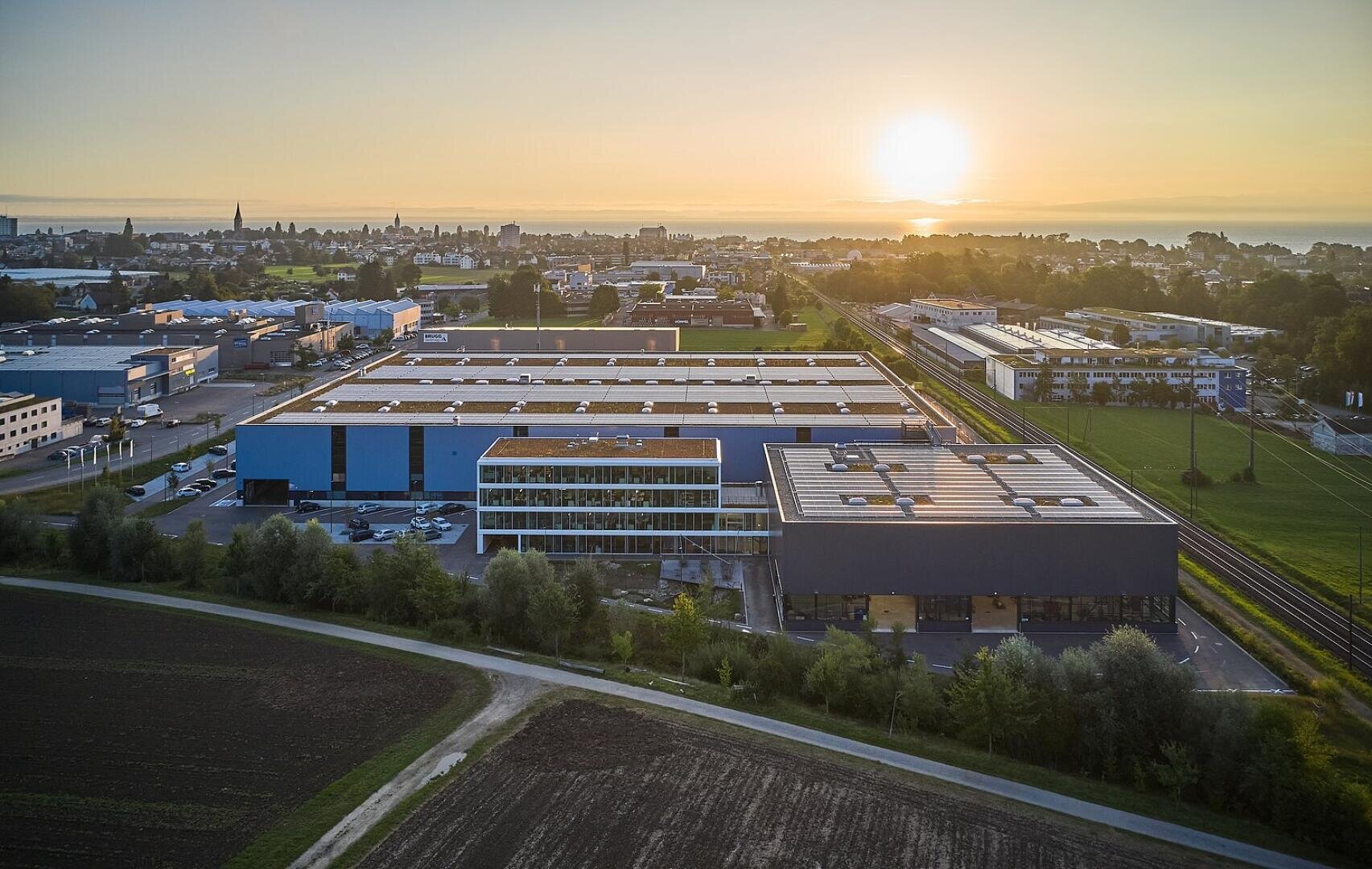 Luftaufnahme von Industriegebäuden und Lagerhallen in einem Gewerbegebiet bei Sonnenuntergang, mit der Skyline der Stadt und offenen Feldern im Hintergrund.