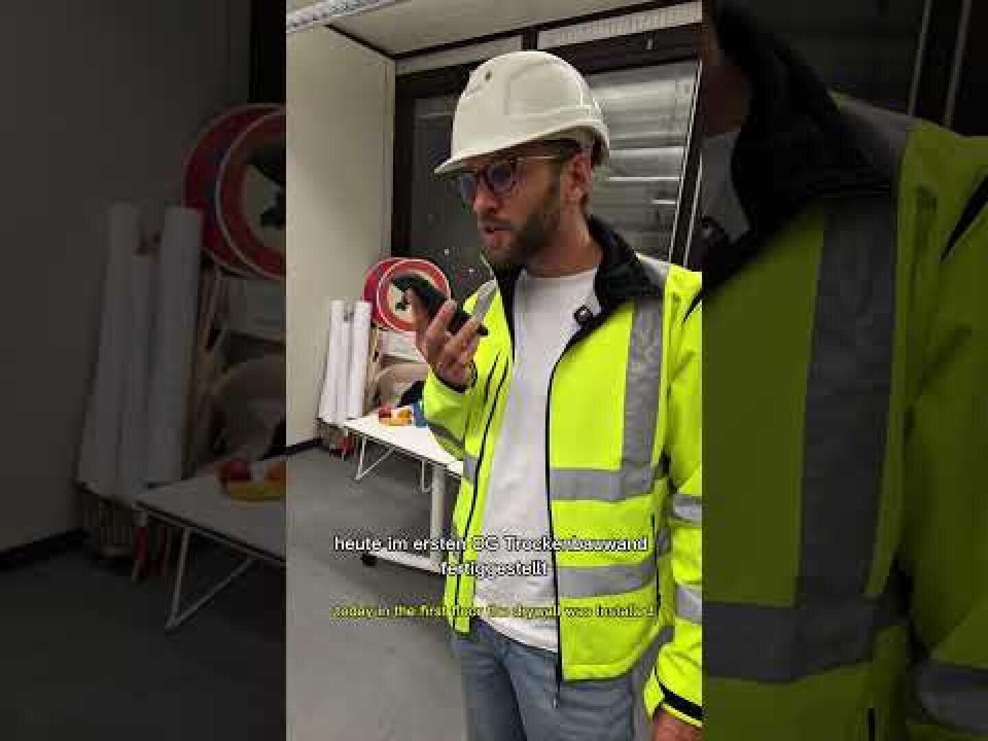 Un homme portant un casque de sécurité blanc, des lunettes et une veste de sécurité jaune vif tient un talkie-walkie dans une main et se tient à l'intérieur, près d'étagères contenant du matériel. Des sous-titres jaunes apparaissent en bas de l'image.