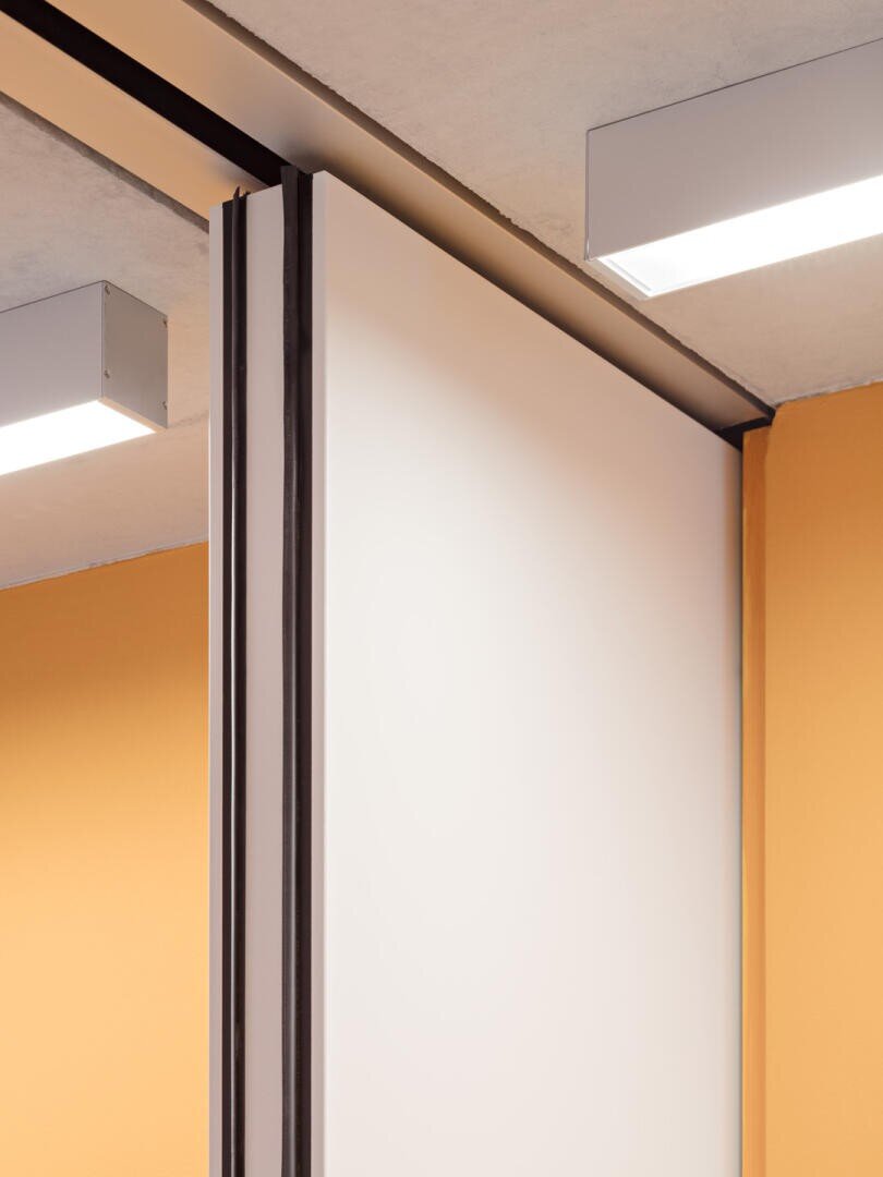 Nahaufnahme einer modernen Schiebetrennwand mit weißem Paneel und schwarzer Zierleiste zwischen gelb-orangenen Wänden und unter einer Betondecke mit rechteckigen Deckenleuchten.