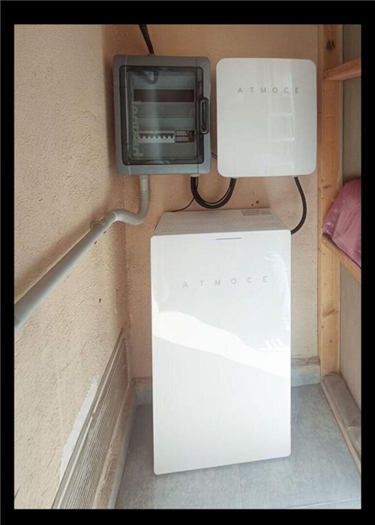 Ein weißes, rechteckiges Gerät mit der Aufschrift ATMOCE steht an einer Wand, darüber sind zwei graue Schaltkästen, ebenfalls mit der Aufschrift ATMOCE, angebracht. Elektrische Leitungen verbinden die Kästen miteinander. Der Bereich ist mit einem Fliesenboden versehen.