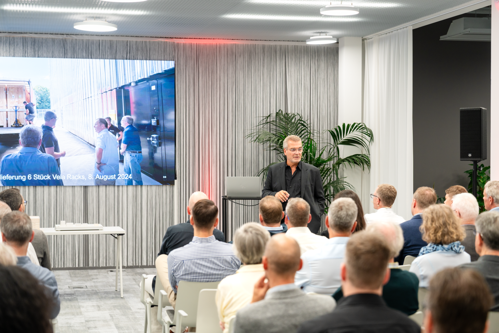 Ein Mann in Schwarz spricht zu einem sitzenden Publikum in einem modernen Konferenzraum. Hinter ihm zeigt ein großer Bildschirm ein Bild von im Freien stehenden Menschen und einen Text in deutscher Sprache. Pflanzen und Vorhänge schmücken den Raum.