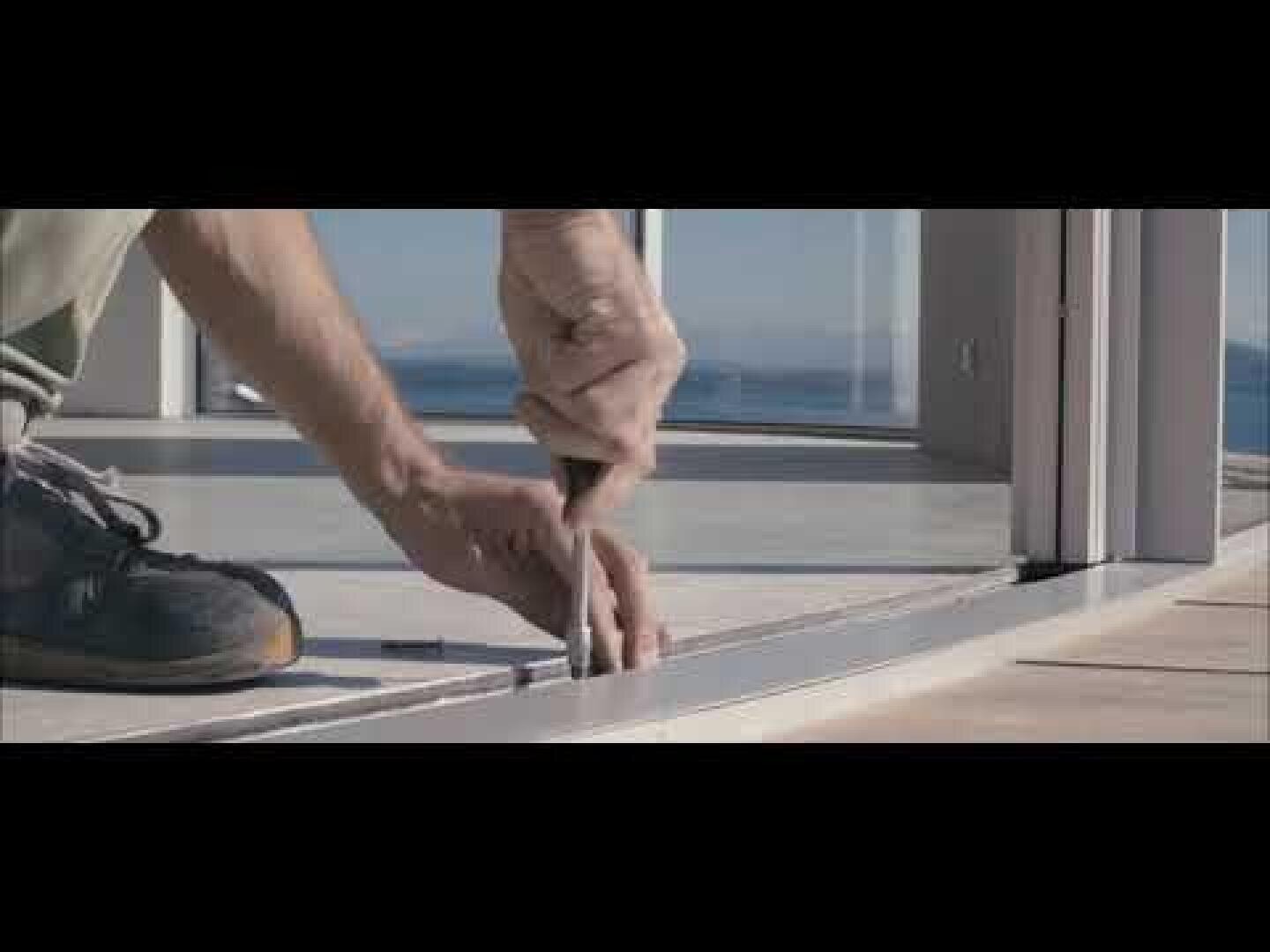 Une personne portant des chaussures de sport utilise un tournevis pour installer ou ajuster le cadre d'une porte coulissante en verre sur un balcon ou une terrasse, avec une vue panoramique en arrière-plan.