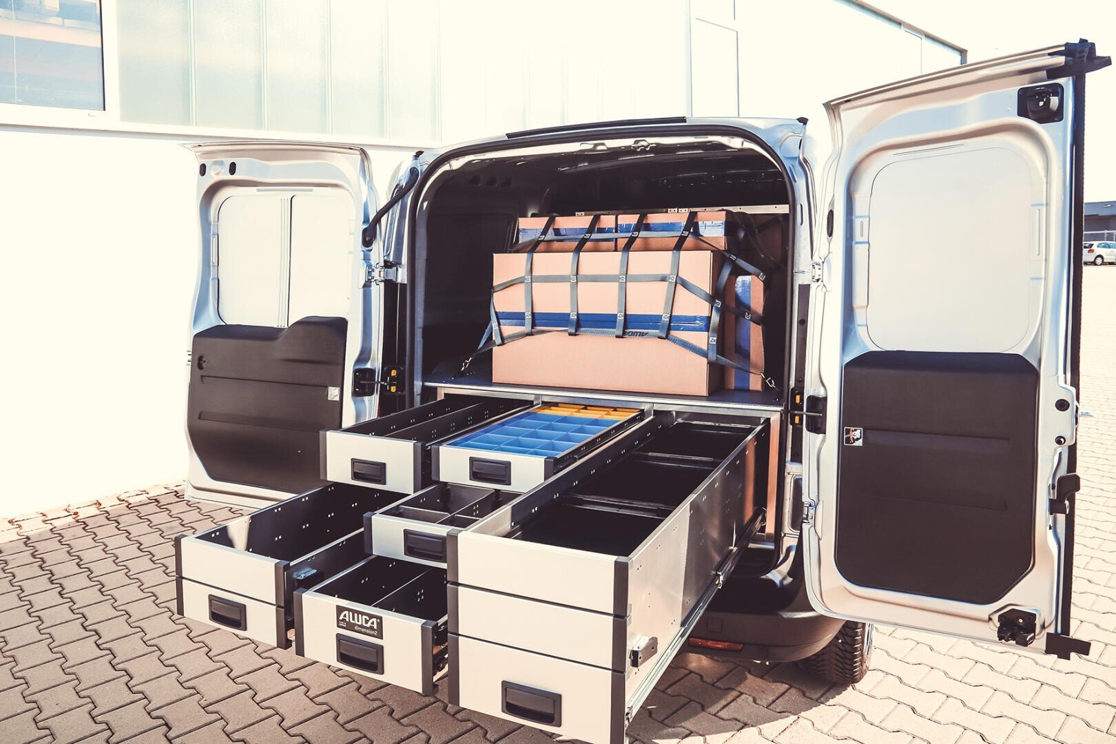 Une camionnette dont les portes arrière sont ouvertes montre des tiroirs de rangement bien ordonnés et des compartiments bleus ainsi qu'une grande caisse sécurisée par des sangles bleues, démontrant ainsi une gestion efficace du chargement.