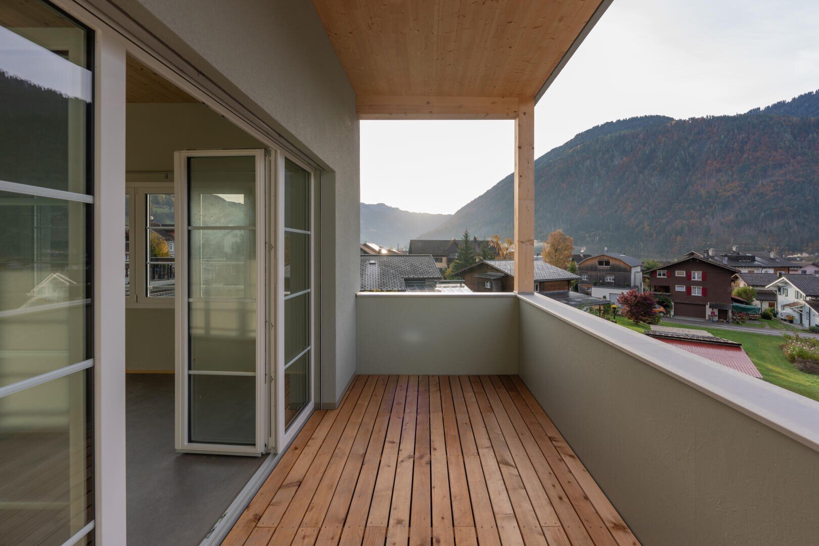 Ein Holzbalkon mit Glastüren bietet einen Blick auf ein kleines Dorf und grüne Hügel mit Bäumen und Bergen in der Ferne, unter einem teilweise bewölkten Himmel bei Sonnenuntergang.