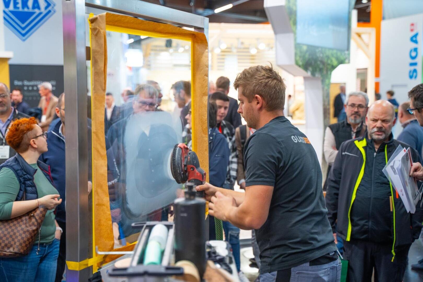 Ein Mann demonstriert ein Elektrowerkzeug auf einer Glasplatte vor einer Menschenmenge auf einer Messe oder Ausstellung. Die Leute schauen aufmerksam zu, und im Hintergrund sind viele Stände und Auslagen zu sehen.