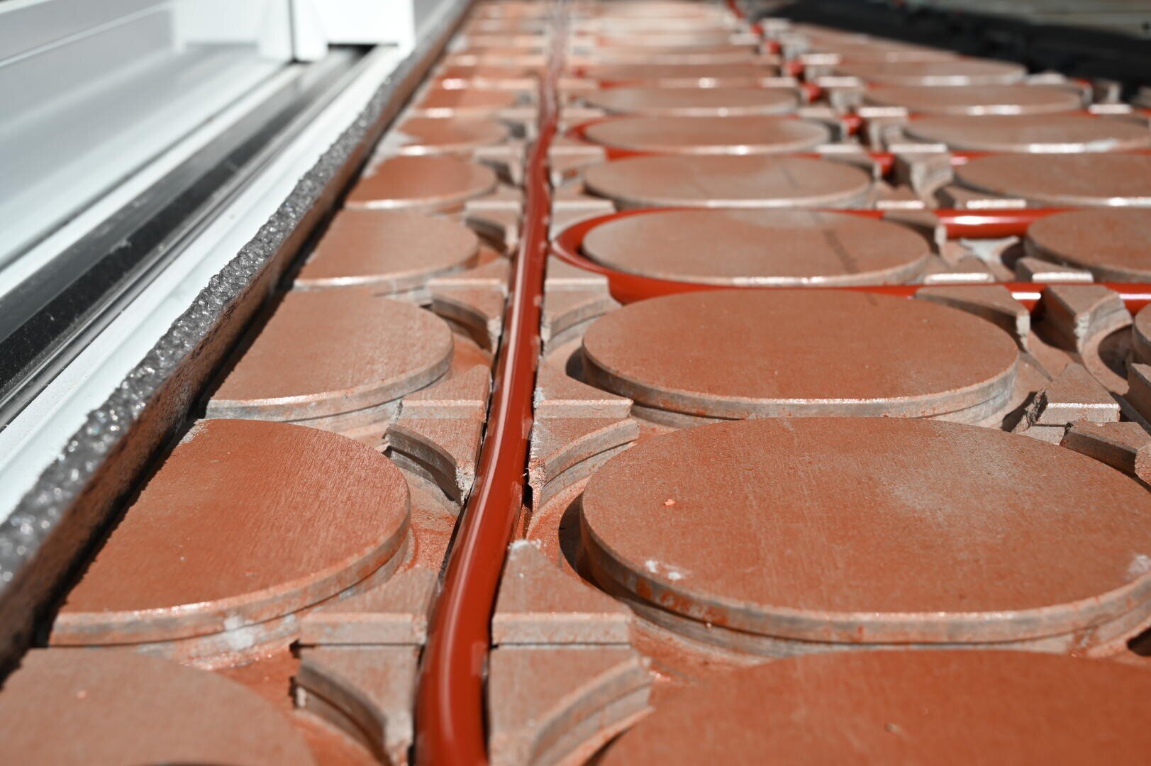 Nahaufnahme eines Fußbodenheizungssystems mit roten Rohren, die in einem Gitter aus runden, braunen Platten in der Nähe einer weißen Wand oder eines Fensterrahmens verlegt sind.