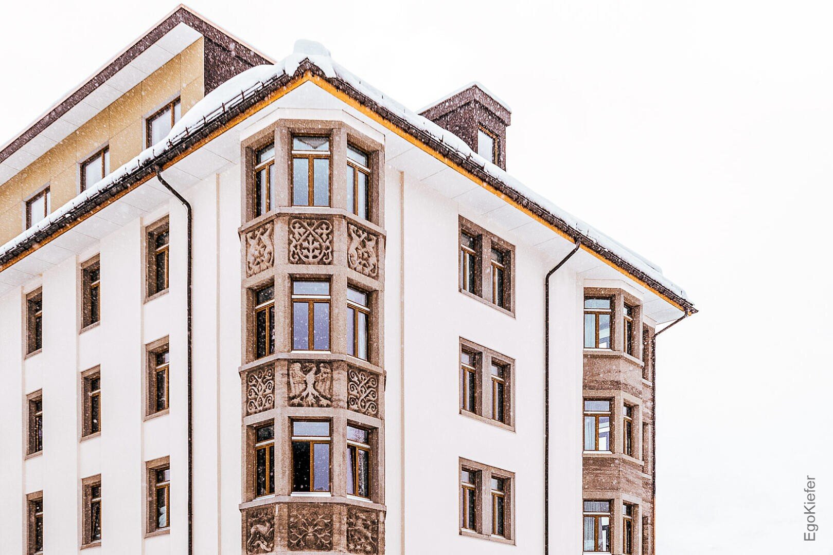 Ein hohes Eckgebäude mit dekorativen Mustern an der Fassade, mehreren großen Fenstern und einem schneebedeckten Dach vor einem hellen, bedeckten Himmel.