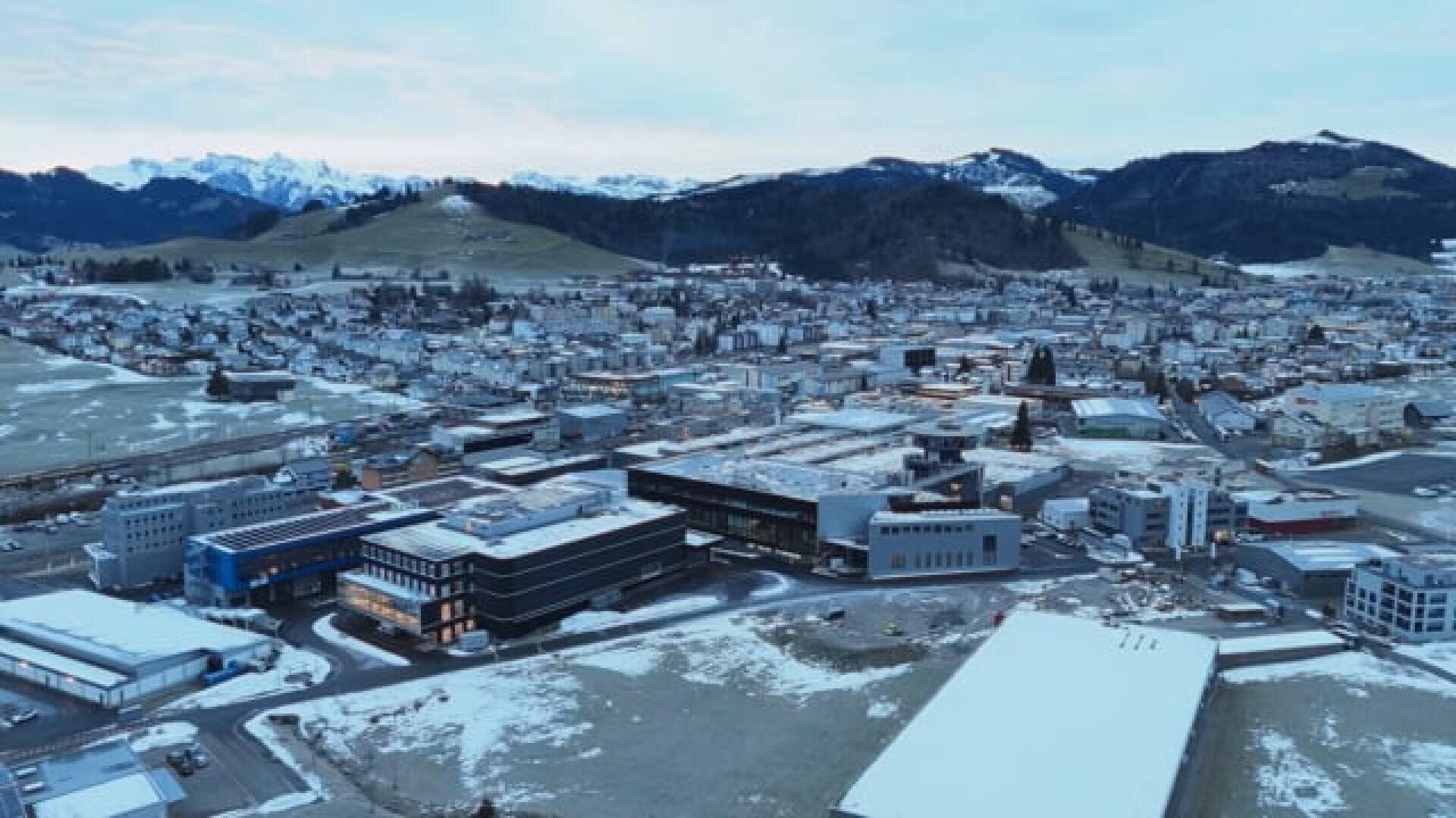 Vue aérienne d'une petite ville avec des bâtiments modernes, une légère neige sur le sol et des collines environnantes. En arrière-plan, on aperçoit des montagnes enneigées sous un ciel nuageux.