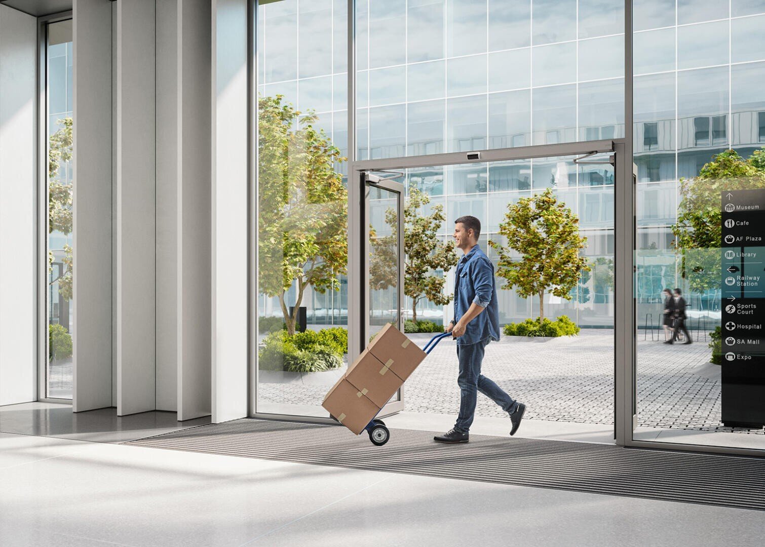 Ein Mann schiebt eine Sackkarre mit einer großen Pappschachtel durch den Glaseingang eines modernen Bürogebäudes, vor dem Bäume und Menschen zu sehen sind.