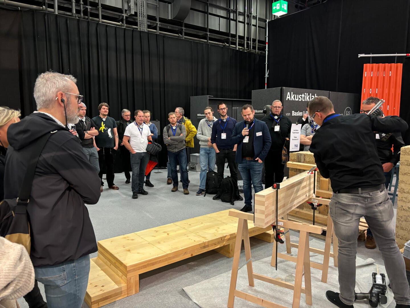 Eine Gruppe von Zuschauern beobachtet eine Live-Vorführung der Holzbearbeitung bei einer Veranstaltung in einer Halle. Zwei Vorführer arbeiten an einer Holzstruktur, während das Publikum steht und aufmerksam zusieht. Die Umgebung ist mit Bänken und Informationsdisplays ausgestattet.