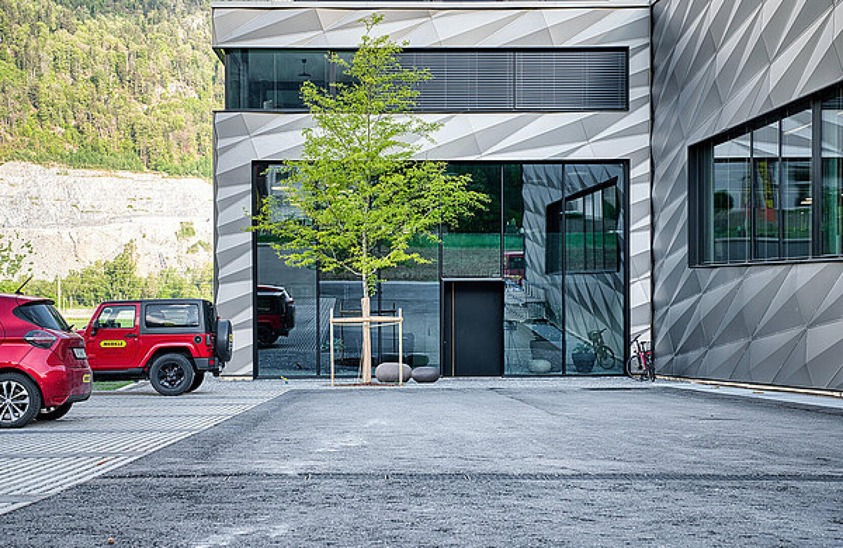 Modernes Gebäude mit geometrischer Fassade, großen Glasfenstern und einem davor gepflanzten jungen Baum. Auf der linken Seite sind ein rotes Auto und ein roter Jeep geparkt, und in der Nähe des Gebäudeeingangs steht ein Fahrrad. Im Hintergrund sind bewaldete Hügel zu sehen.