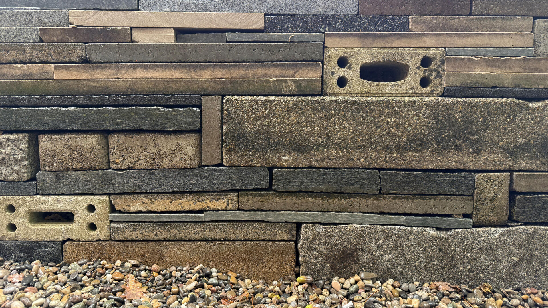 Nahaufnahme einer Steinmauer aus rechteckigen und unregelmäßig geformten Ziegeln in verschiedenen Grau- und Brauntönen, wobei einige Ziegel Löcher aufweisen. Kleine Kieselsteine bedecken den Boden am Fuß der Mauer.