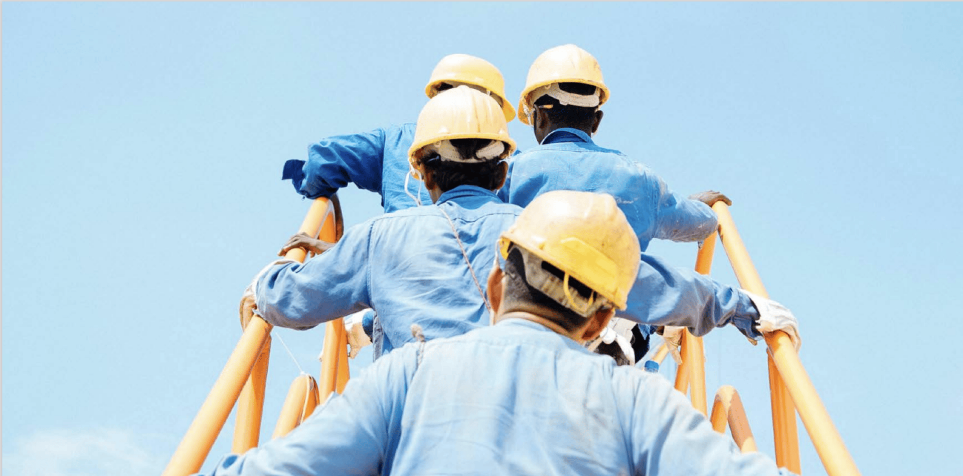 Vier Arbeiter in blauen Uniformen und gelben Sicherheitshelmen klettern an einer orangefarbenen Metallkonstruktion hoch, die sich vor einem strahlend blauen Himmel befindet. Das Foto ist von hinten aufgenommen und zeigt Teamwork und Arbeitssicherheit.