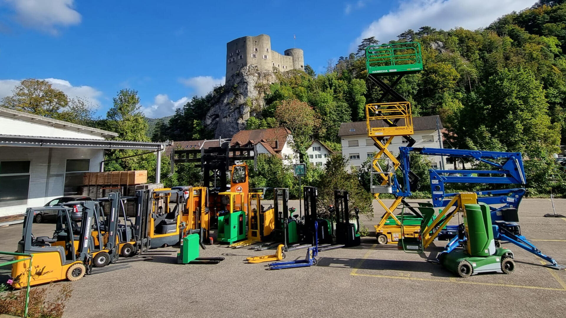 Verschiedene Gabelstapler und Hebegeräte sind auf einem gepflasterten Parkplatz abgestellt. Im Hintergrund sind Häuser, Bäume, ein felsiger Hügel und eine große Steinburg bei teilweise bewölktem Himmel zu sehen.