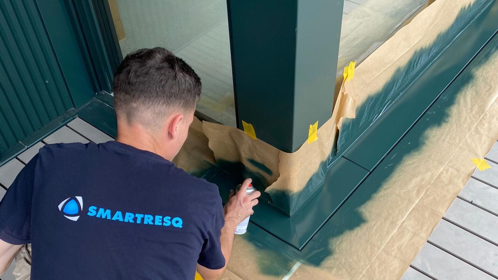 Eine Person, die ein SMARTRESQ-Hemd trägt, besprüht eine grüne Metallsäule, wobei Abdeckpapier und Klebeband den umliegenden Bereich und den Boden schützen.