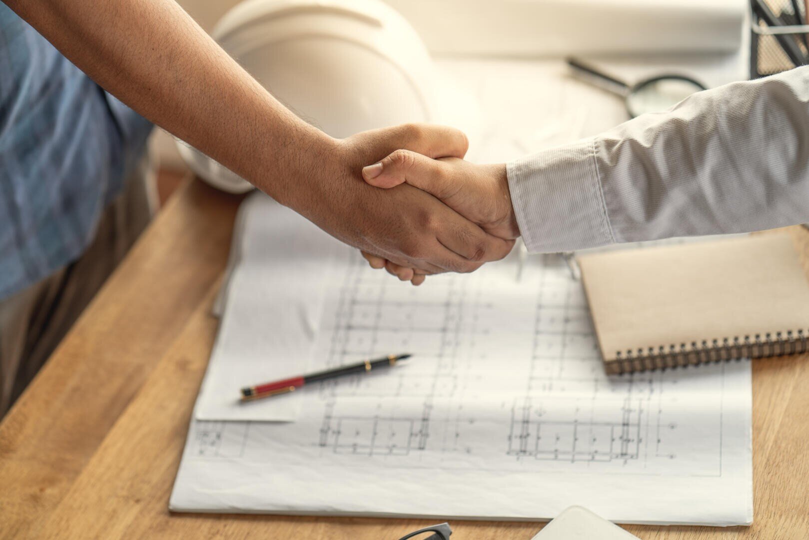 Deux personnes se serrent la main au-dessus d'un bureau contenant des plans architecturaux, un stylo, un carnet de notes et un casque de protection blanc, ce qui indique un accord professionnel ou une collaboration dans le domaine de la construction ou de l'ingénierie.