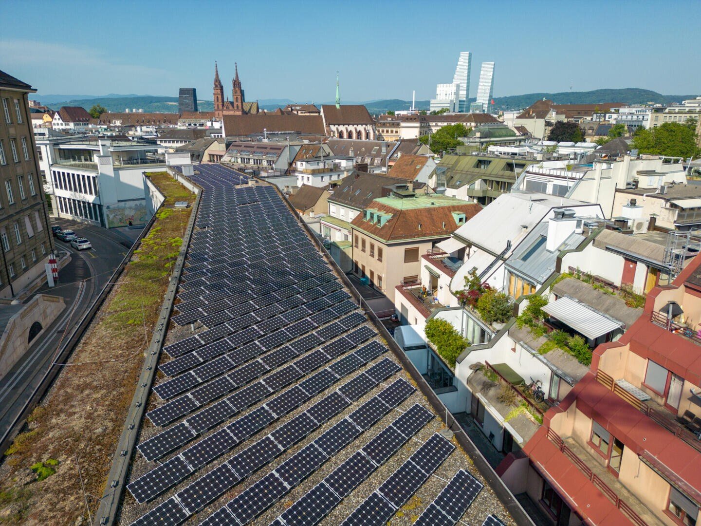 Luftaufnahme einer Stadt mit Sonnenkollektoren auf einem großen, flachen Dach. Umliegende Gebäude und historische Kirchen sind zu sehen, zusammen mit modernen Hochhäusern im Hintergrund unter einem klaren Himmel.