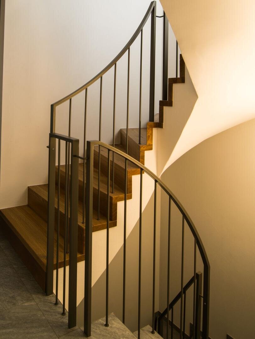 La lumière du soleil éclaire un escalier moderne en bois avec des rampes en métal noir, projetant des ombres distinctives sur les murs blancs et soulignant le design incurvé de l'escalier.