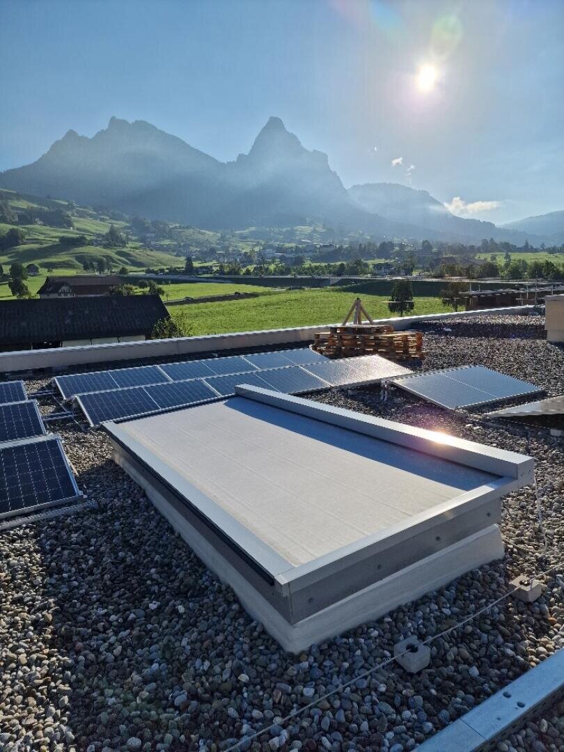 Des panneaux solaires sur un toit de gravier avec des montagnes et un ciel ensoleillé en arrière-plan, des champs verdoyants et un petit village au loin.