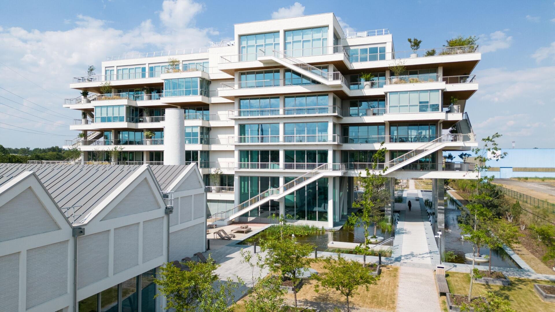 Il s'agit d'un immeuble moderne de plusieurs étages avec de grandes baies vitrées et des terrasses, doté d'escaliers extérieurs blancs. Dans l'espace vert devant le bâtiment se trouvent des arbres et un trottoir, à gauche duquel se trouve un autre bâtiment.