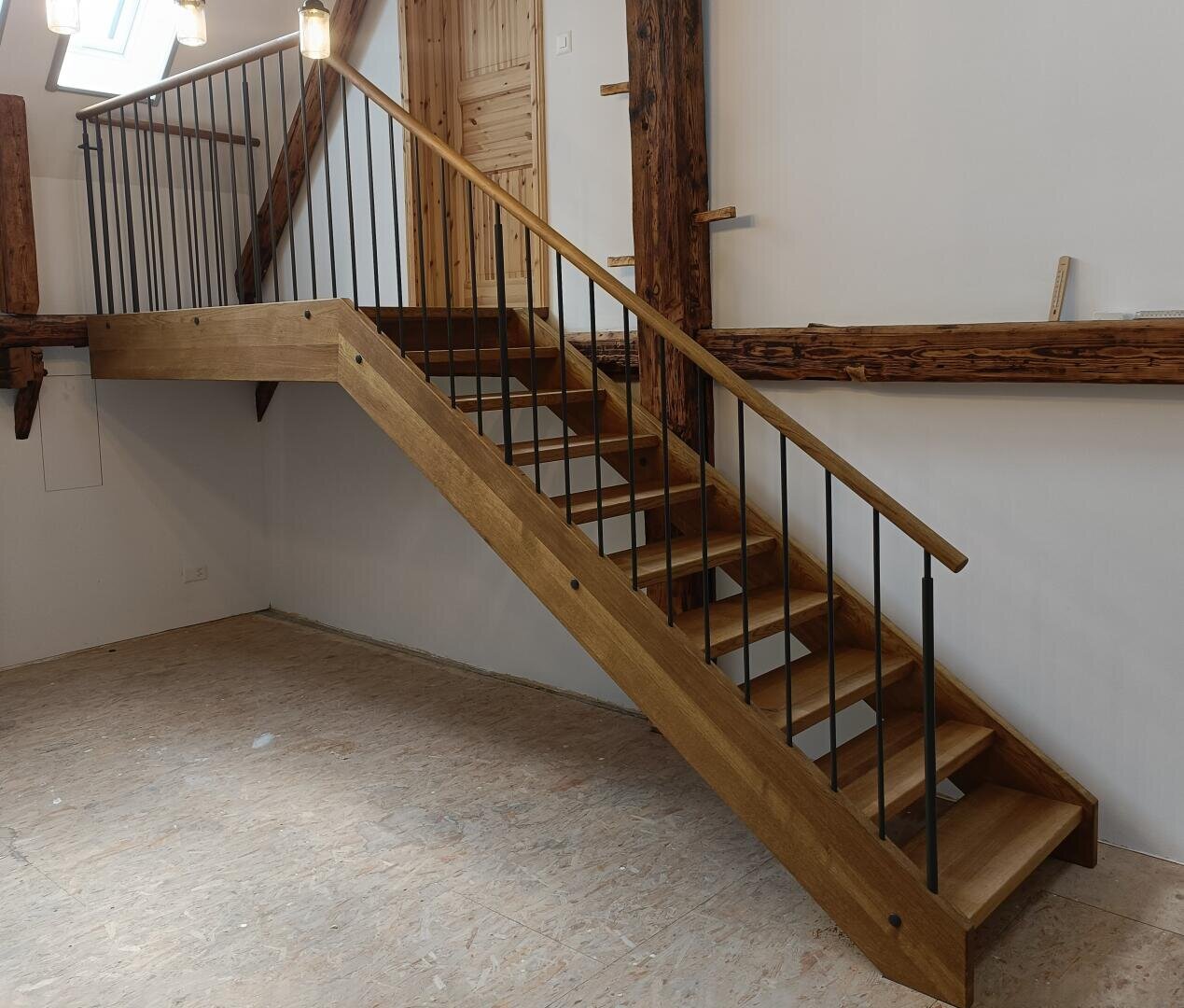 Un escalier en bois avec une rampe en métal mène à un étage supérieur dans une pièce au sol brut et aux poutres en bois exposées sur les murs. La pièce semble être en cours de rénovation.