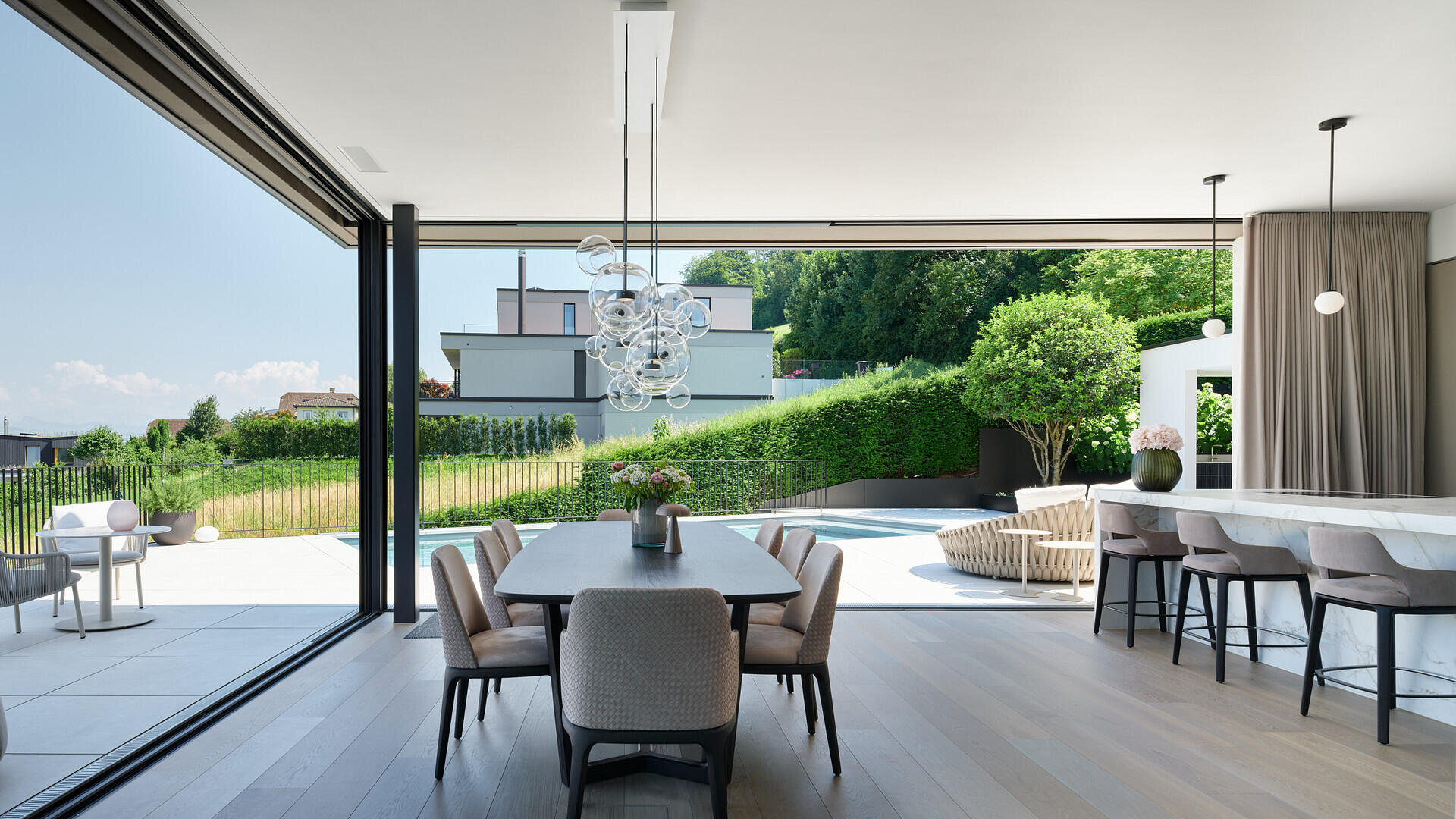 Une salle à manger et une cuisine modernes et ouvertes avec de grandes portes coulissantes en verre qui donnent sur une terrasse avec des sièges en plein air, une piscine et une cour verdoyante avec des bâtiments modernes en arrière-plan.