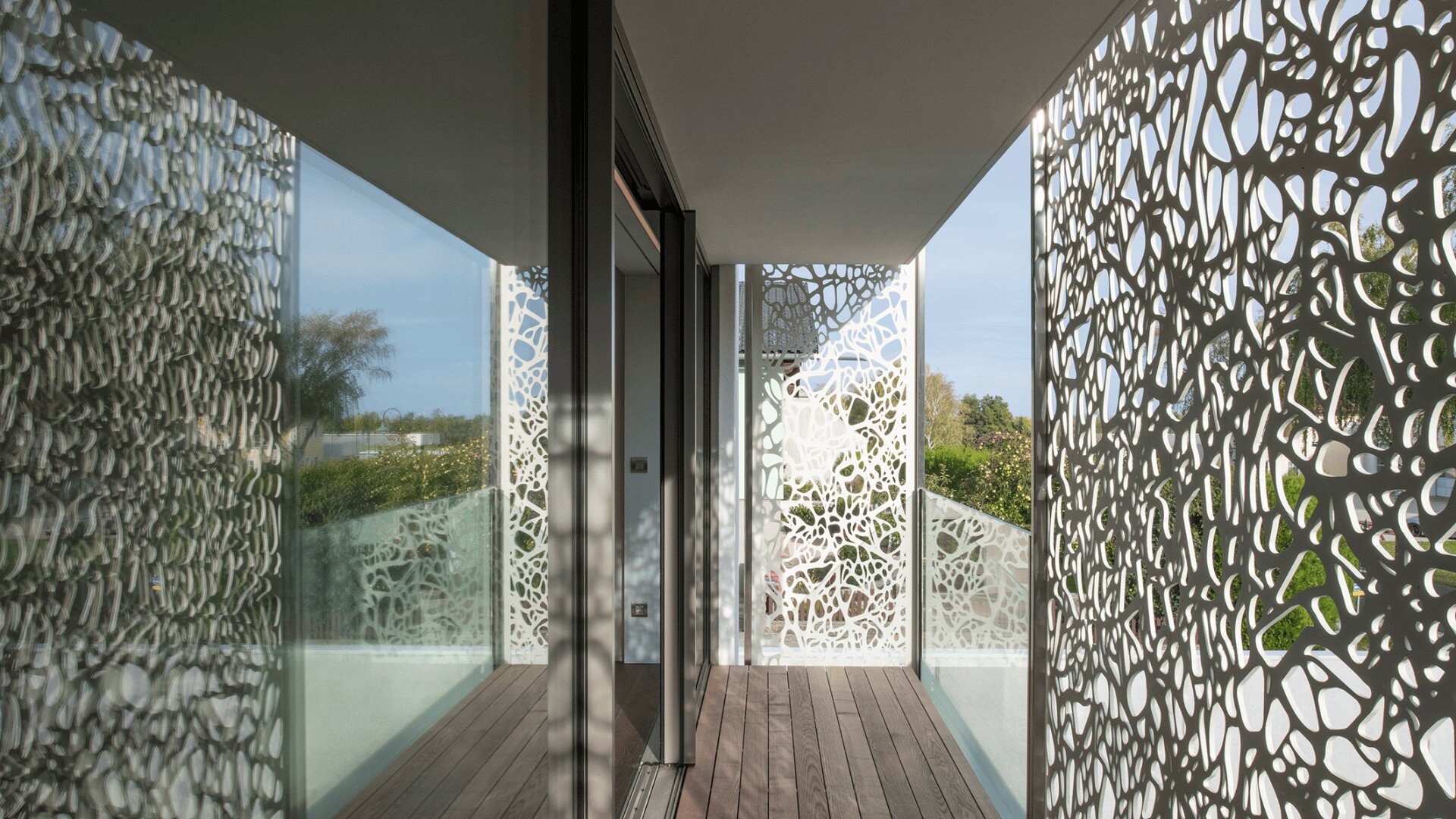 Ein moderner Balkon mit Holzfußboden und Glaswänden, der mit dekorativen weißen Metallpaneelen mit abstrakten Ausschnitten versehen ist, die komplizierte Schatten auf den Boden und die Wände werfen.