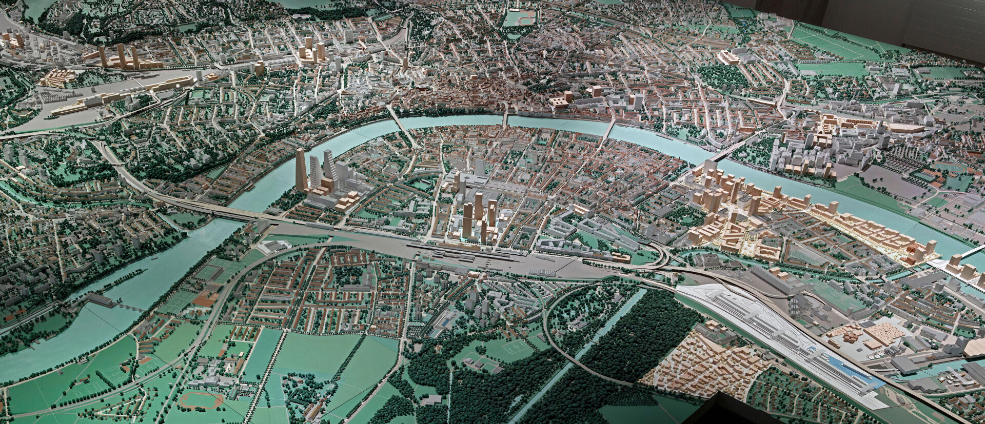 Ein detailliertes, maßstabsgetreues Modell einer Stadt, durch die ein Fluss fließt, umgeben von Gebäuden, Grünflächen, Straßen und Brücken, das die Stadt- und Wohngebiete aus der Luftperspektive zeigt.