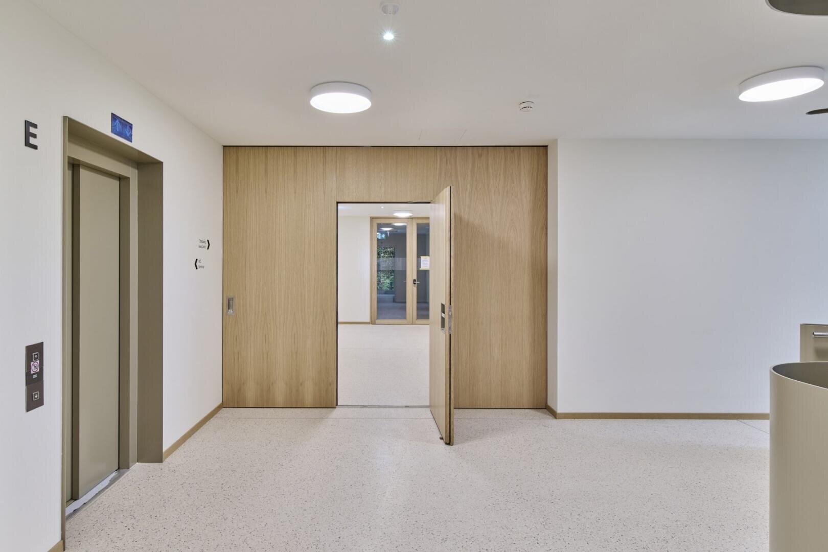 Ein moderner Flur mit hellen Wänden und Fußböden, ein Aufzug auf der linken Seite mit der Aufschrift E und doppelte Holztüren in der Mitte, die zu einem weiteren Korridor mit einem Fenster am Ende führen.