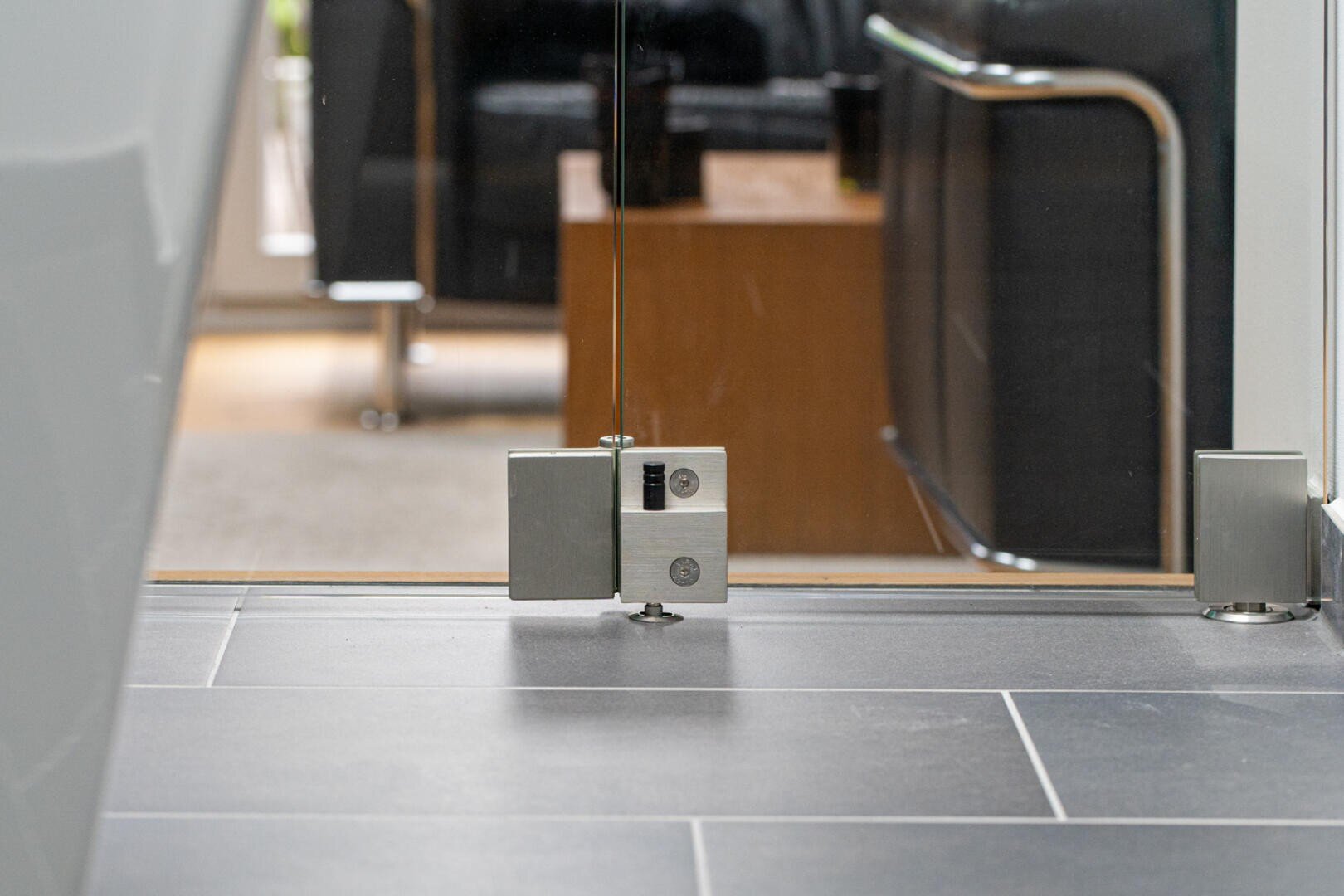 Nahaufnahme eines Türstoppers aus Metall und eines Scharniermechanismus, der an der Unterseite einer Glastür befestigt ist, die auf einem grauen Fliesenboden steht, wobei im Hintergrund moderne Möbel zu sehen sind.