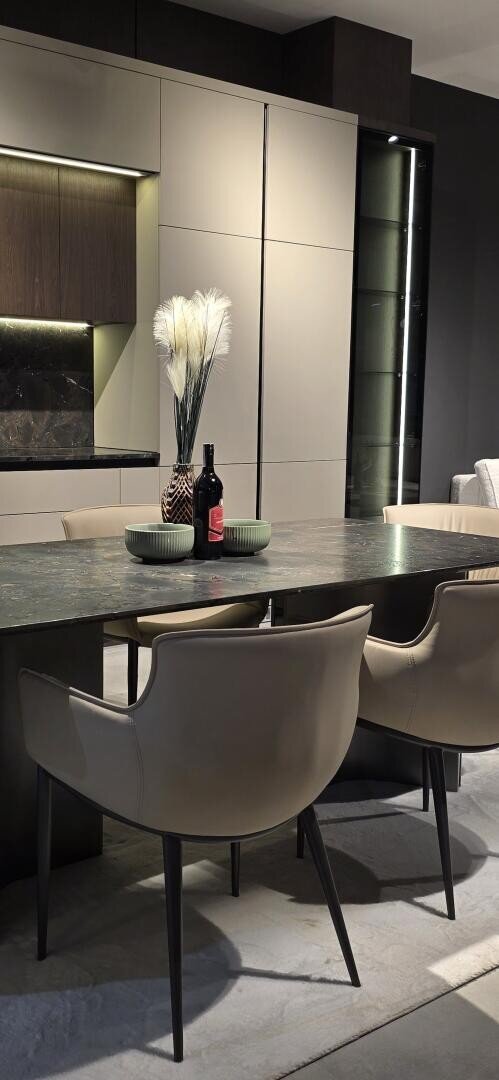 Une salle à manger moderne avec une table en pierre foncée, des chaises beiges, un bol vert, une bouteille de vin rouge et un vase avec des herbes séchées blanches décoratives, le tout entouré d'une armoire élégante et d'un éclairage d'appoint.