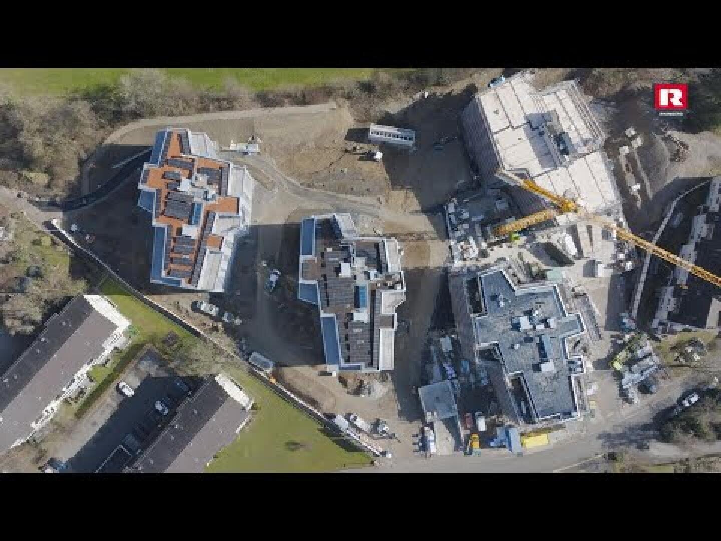 Vue aérienne d'un chantier de construction avec plusieurs bâtiments à différents stades d'achèvement, entourés de terre, de machines et d'arbres. Sur le côté droit de l'image, on peut voir une grue jaune.