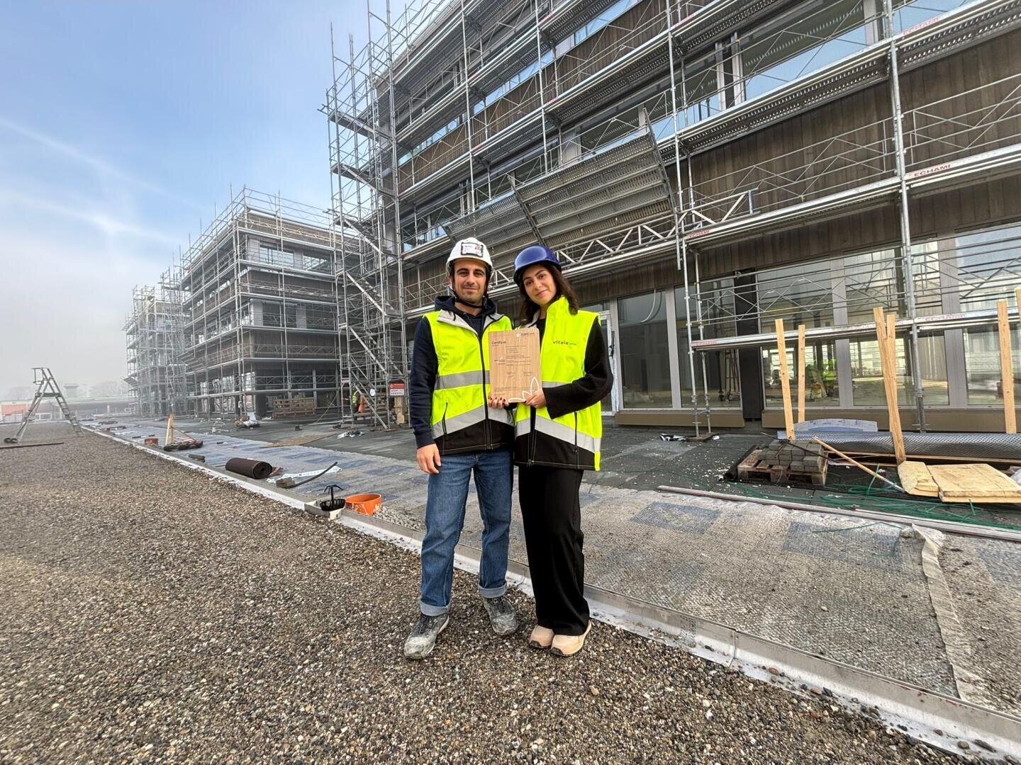 Deux personnes portant des gilets de sécurité et des casques de protection se tiennent sur un chantier de construction devant un bâtiment moderne doté d'un échafaudage. L'une d'elles tient une planchette à pince et toutes deux sourient à la caméra. Le sol est caillouteux et le ciel est dégagé.