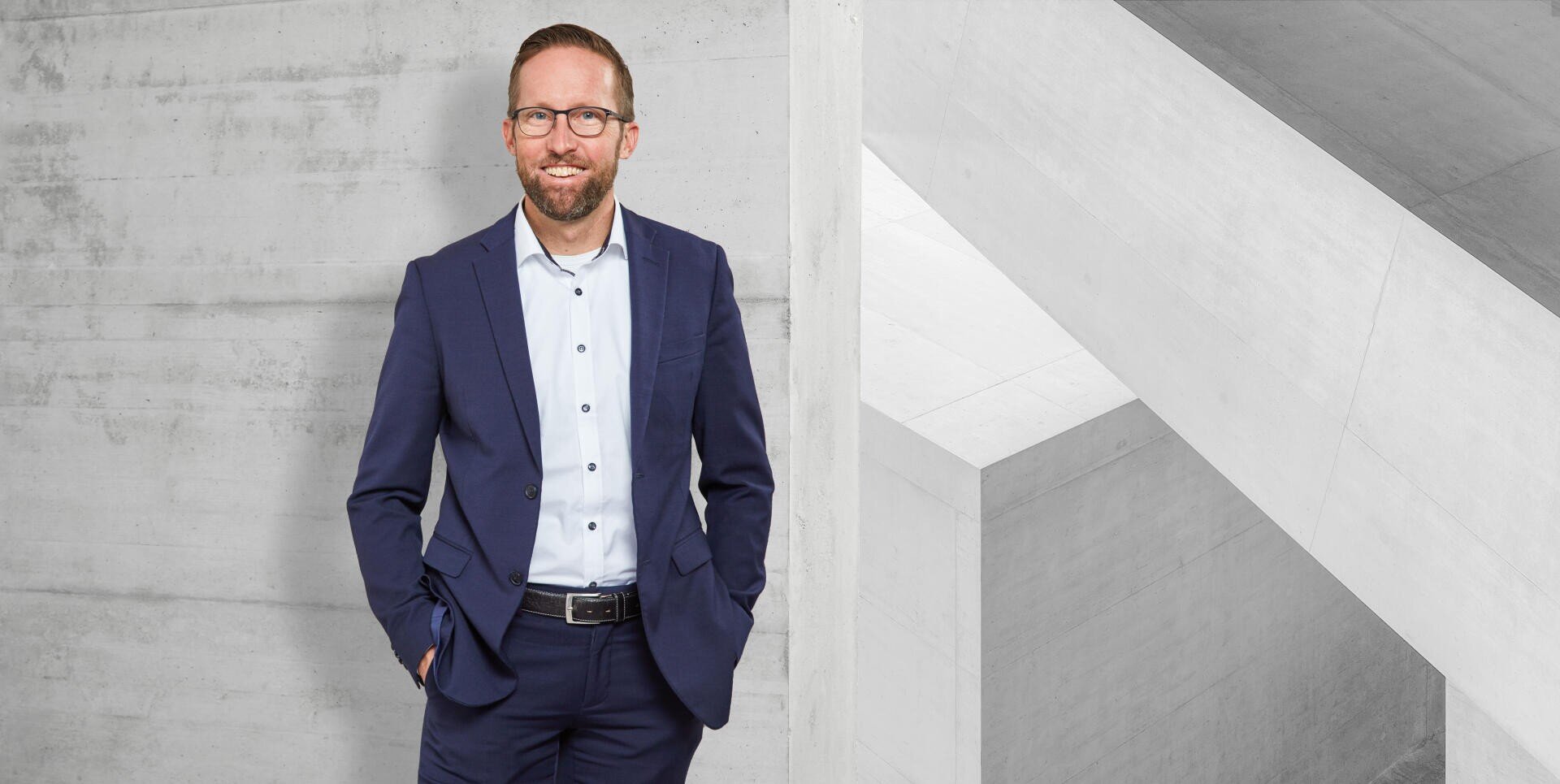 Ein Mann mit Brille und blauem Anzug steht lächelnd mit einer Hand in der Tasche vor dem Hintergrund eines modernen Betontreppenhauses.